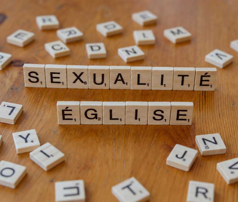les mots Sexualité et Église formés par des lettres de Scrabble en bois sur une table parmi d'autres lettres éparpillées.